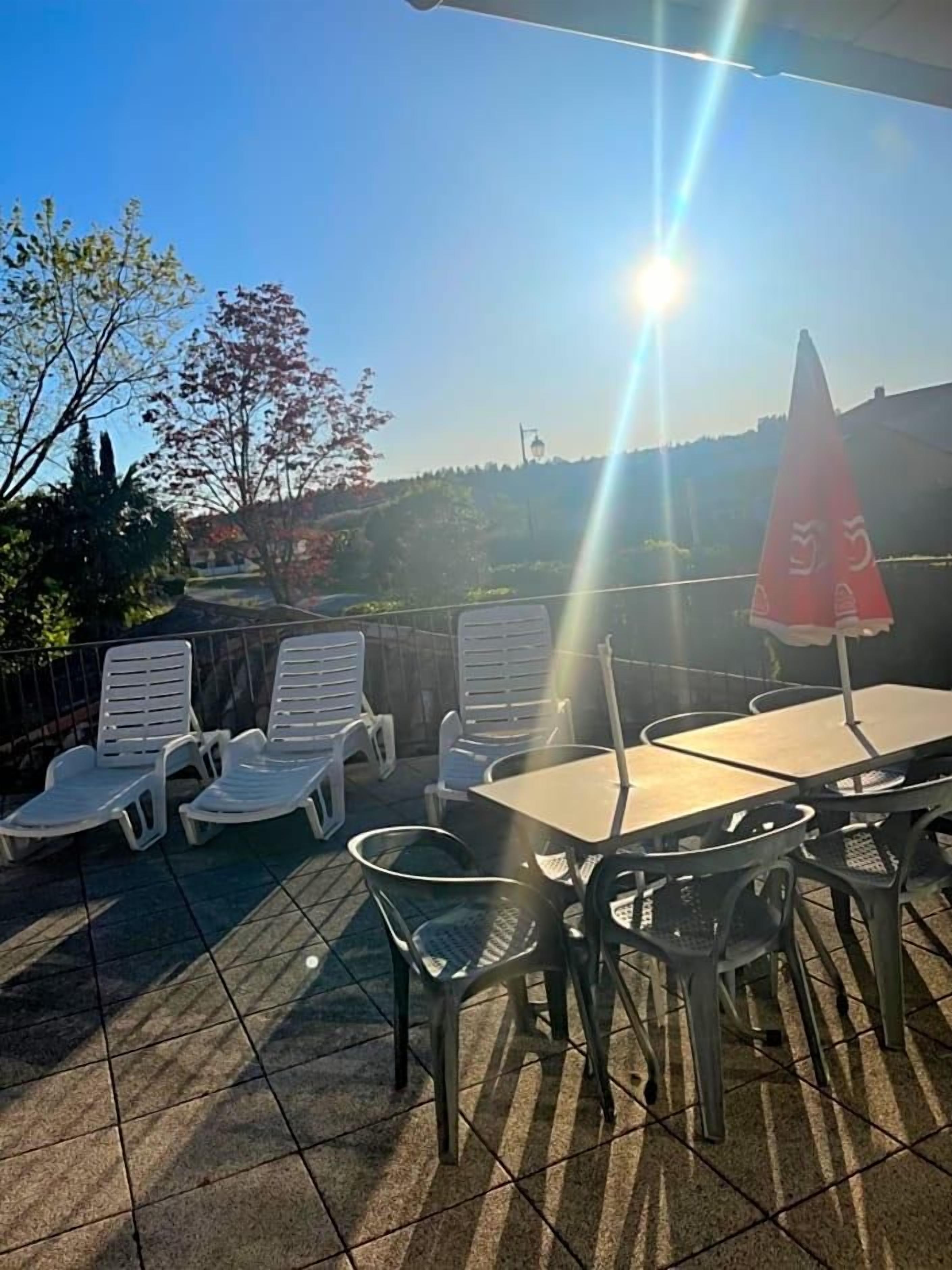 Ein Außenbereich mit Liegestühlen und einem Tisch, einem roten Sonnenschirm und Blick auf Bäume und eine entfernte Landschaft.