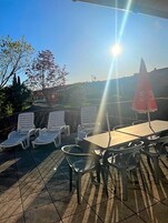 Un área de estar al aire libre con sillones de descanso y una mesa, un paraguas rojo y vistas a árboles y un paisaje lejano.