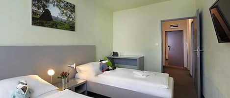 Habitación de hotel con dos camas individuales, una mesita de noche con lámpara, un escritorio pequeño con silla, un cuadro en la pared y una puerta que da a otra habitación.