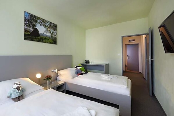 Habitación de hotel con dos camas individuales, una mesita de noche con lámpara, un escritorio pequeño con silla, un cuadro en la pared y una puerta que da a otra habitación.