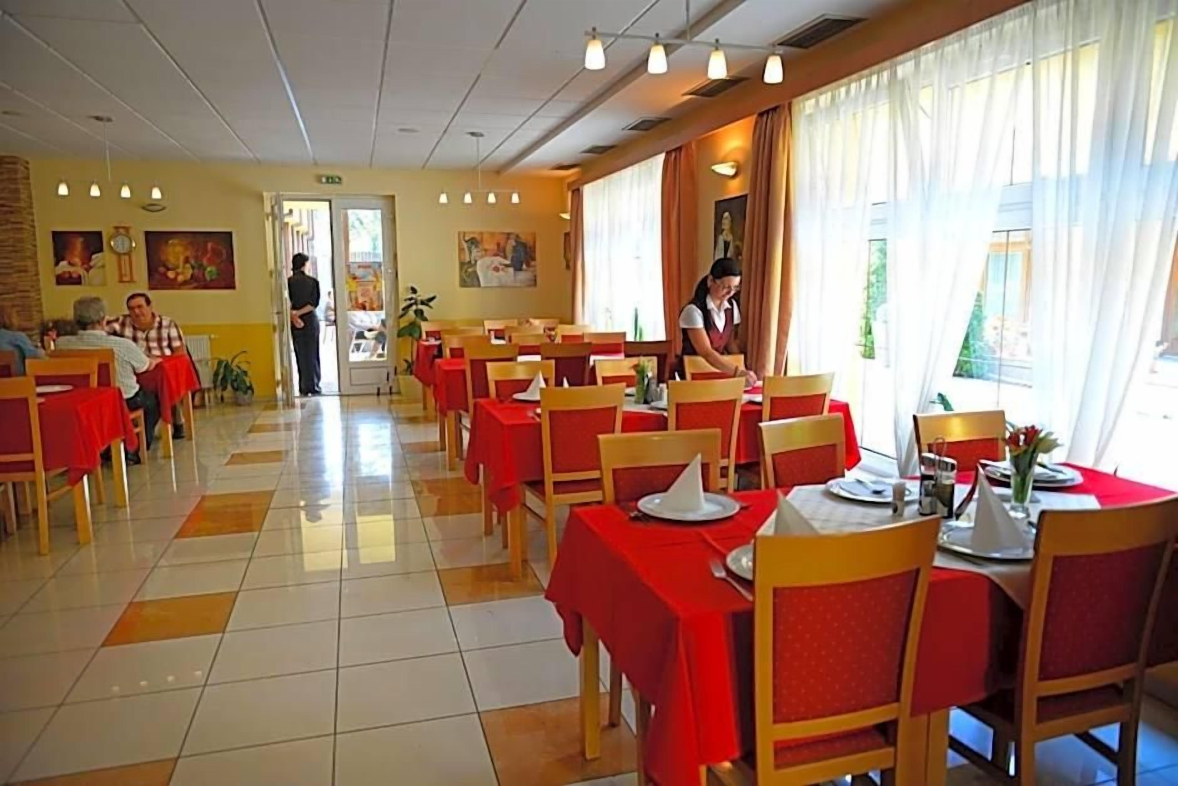 A dining area with tables set for meals, a person serving, and a view of the outdoors through large windows.