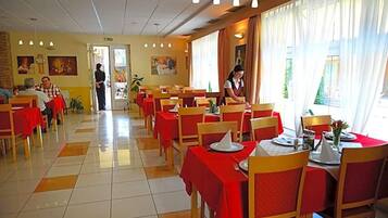 A dining area with tables set for meals, a person serving, and a view of the outdoors through large windows.