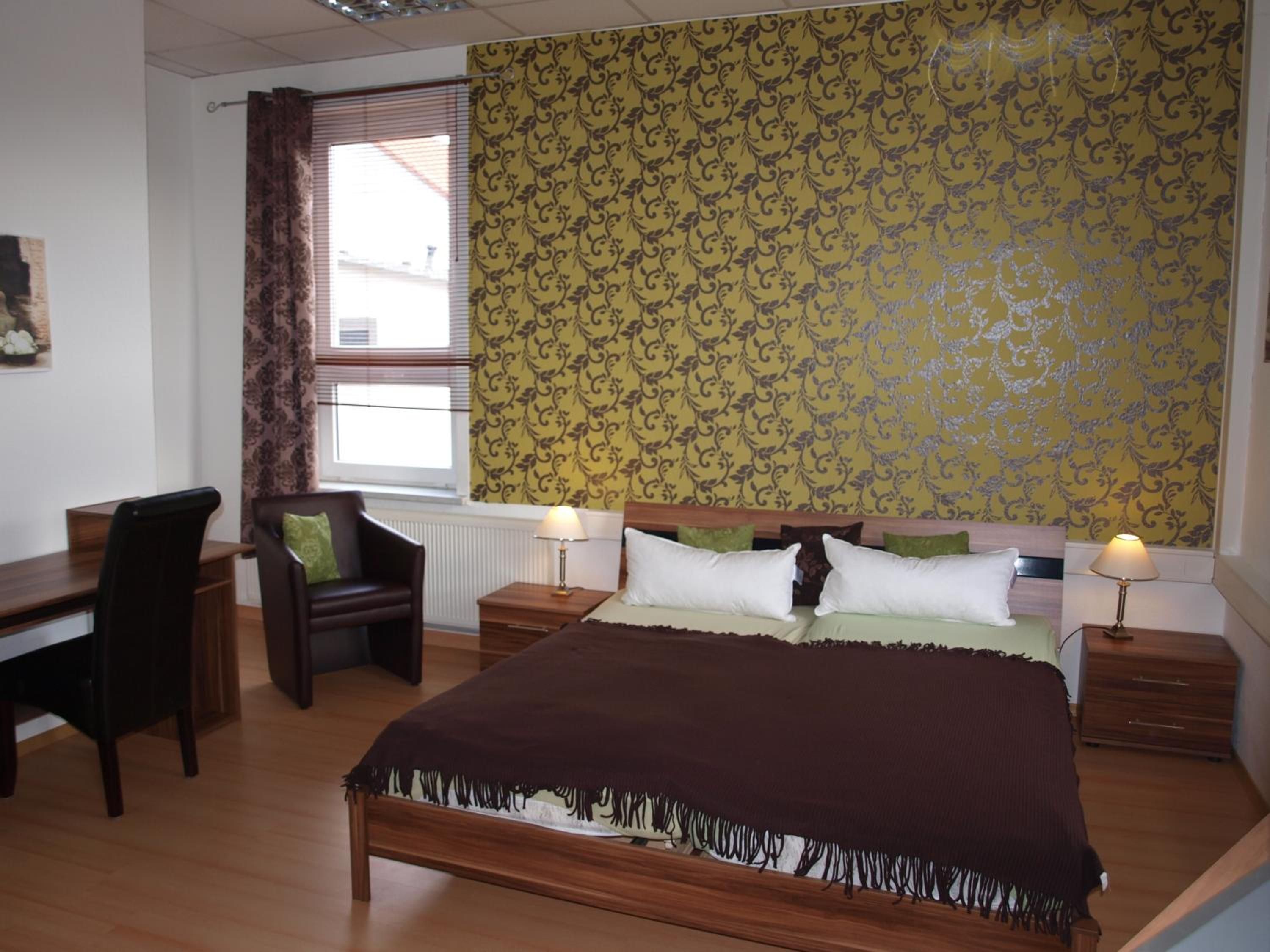 Un dormitorio con cama, mesitas de noche, una silla, escritorio y ventana con cortinas.