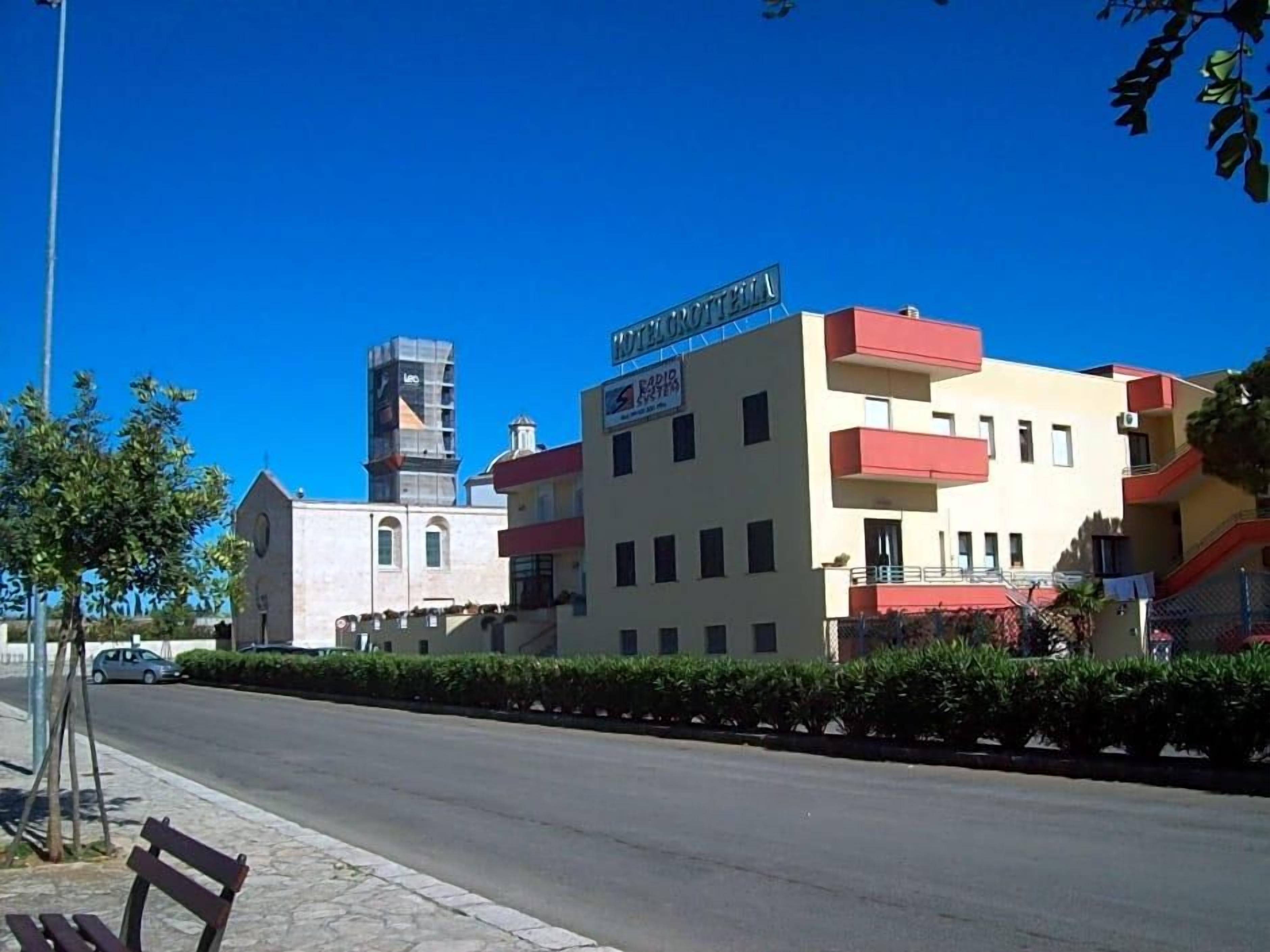 Un hotel llamado 'HOTEL GROTTELE' con una fachada moderna, un estacionamiento visible y un cielo despejado.