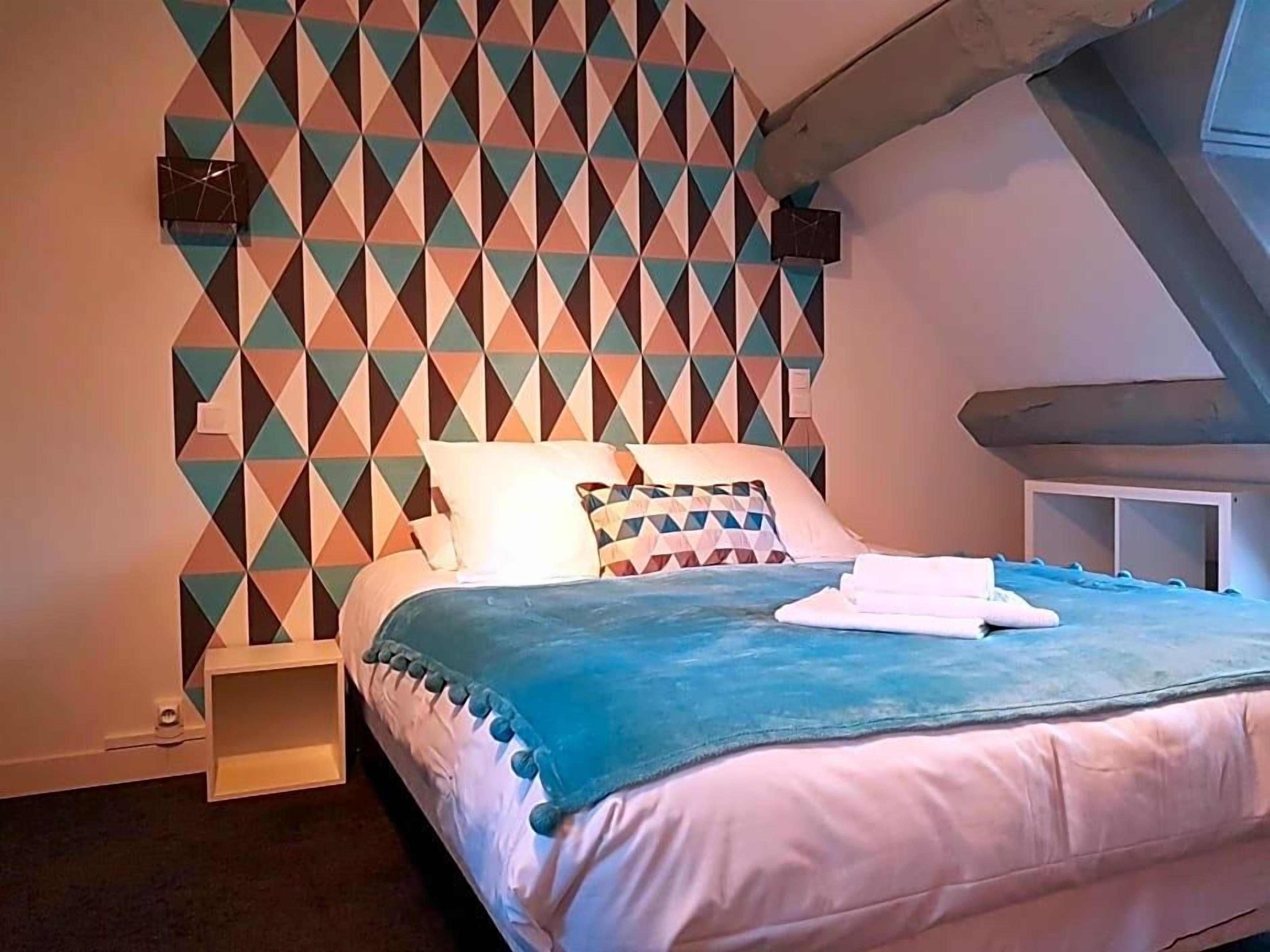 Une chambre avec un mur orné d’un motif géométrique, un lit recouvert d’une couverture bleue et un oreiller blanc.