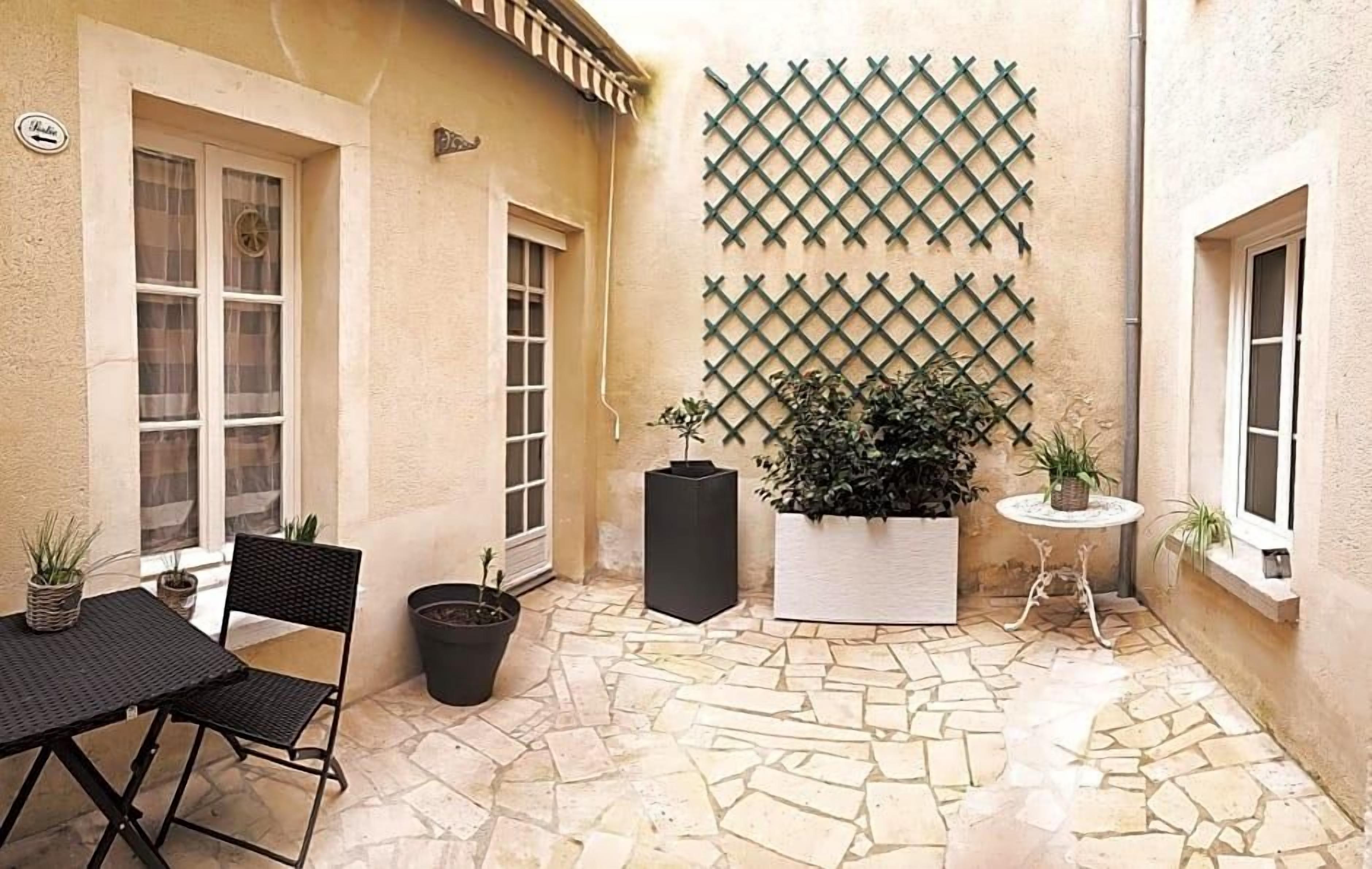 Pátio com piso de pedra, mesa e cadeira de metal preto, plantas em vasos e um muro treliçado.