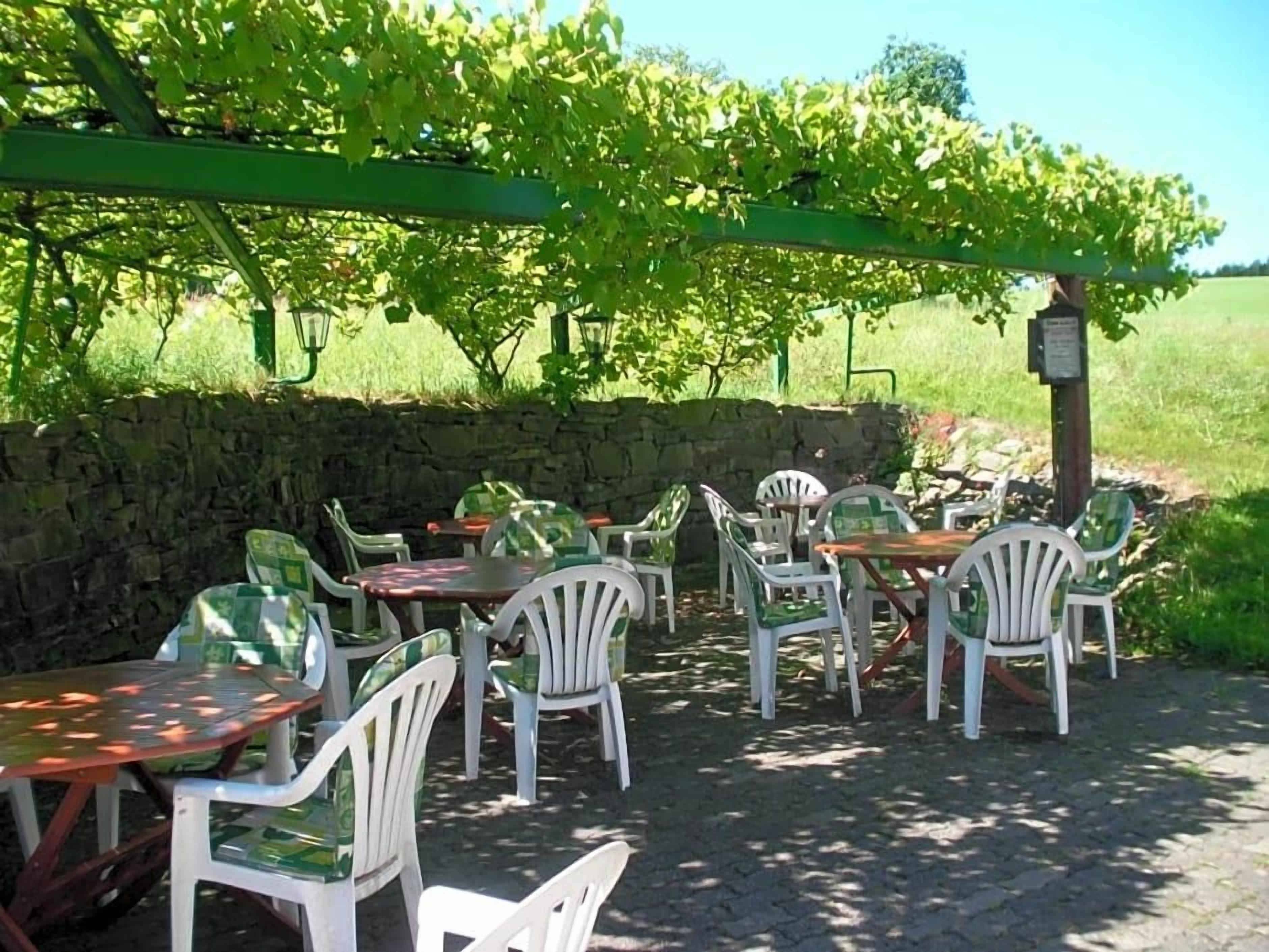 An outdoor seating area with tables and chairs under a pergola, surrounded by a stone wall and a vineyard in the background.