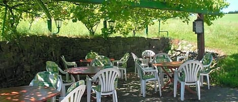 An outdoor seating area with tables and chairs under a pergola, surrounded by a stone wall and a vineyard in the background.