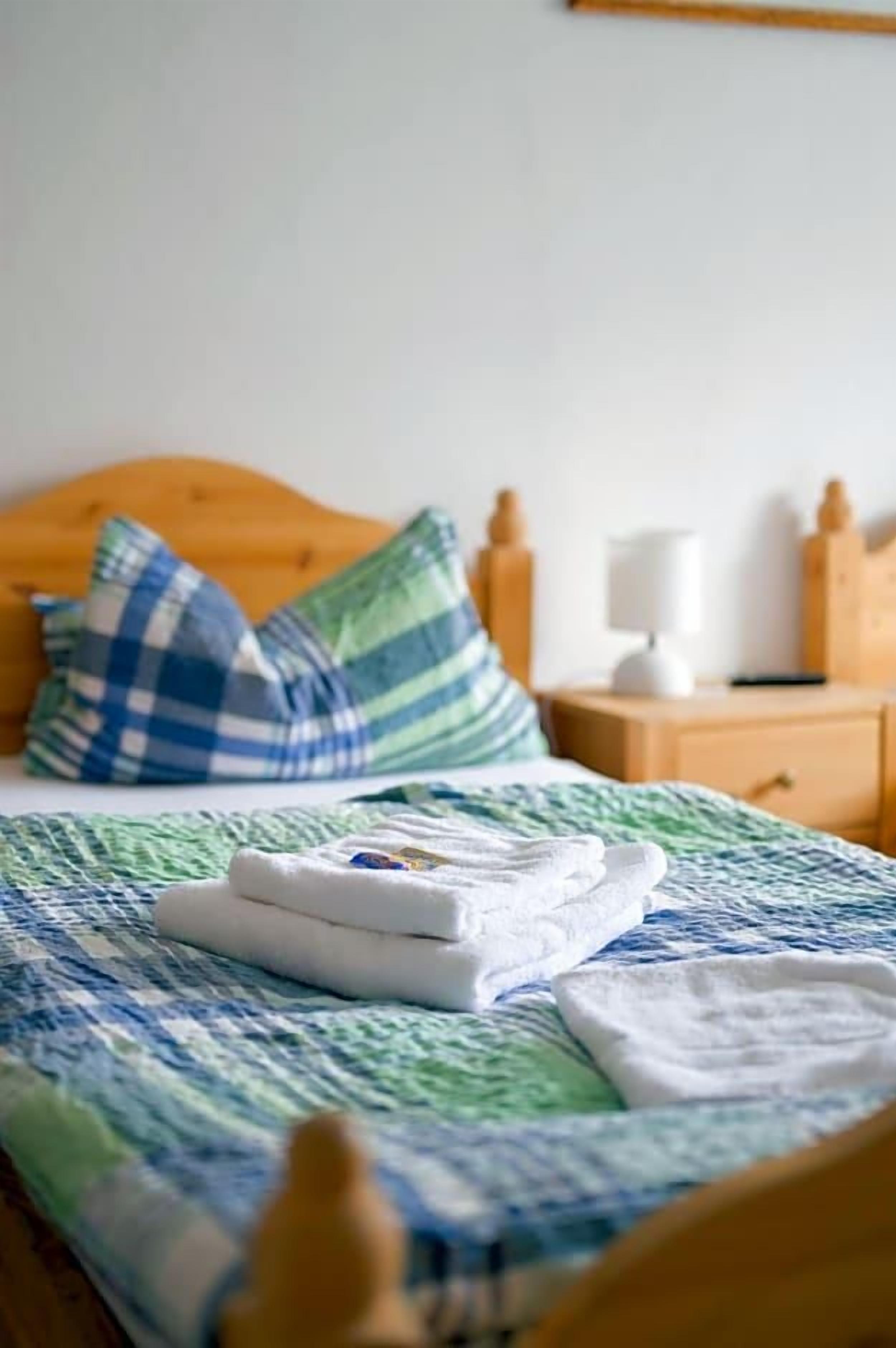 Una cama bien hecha con colcha azul y verde estampada, dos almohadas y una toalla blanca doblada.