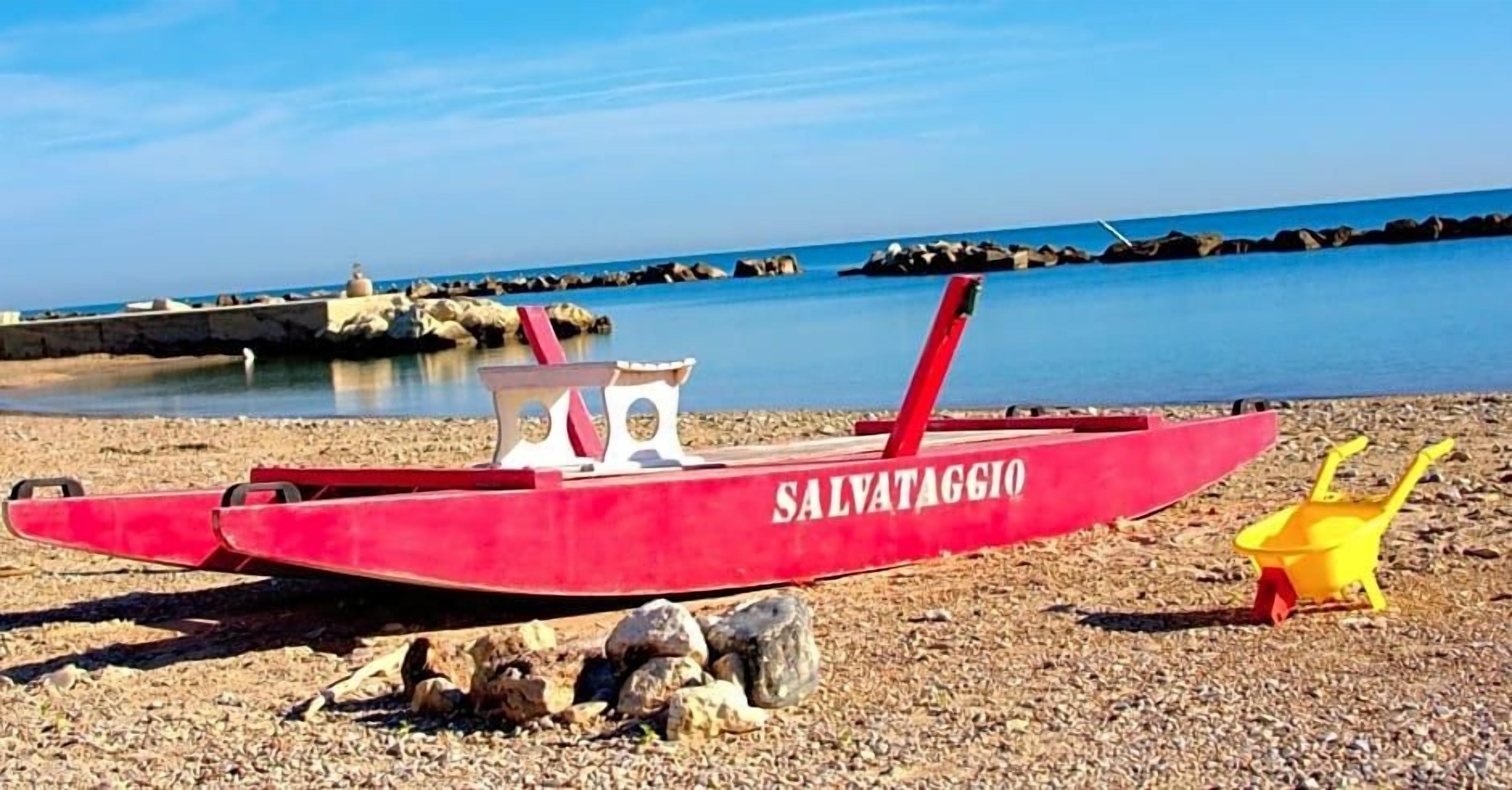 Un bateau rouge portant l’inscription « SALVATAGGIO » était amarré sur une plage de galets, avec un ciel dégagé et une mer calme en arrière-plan.