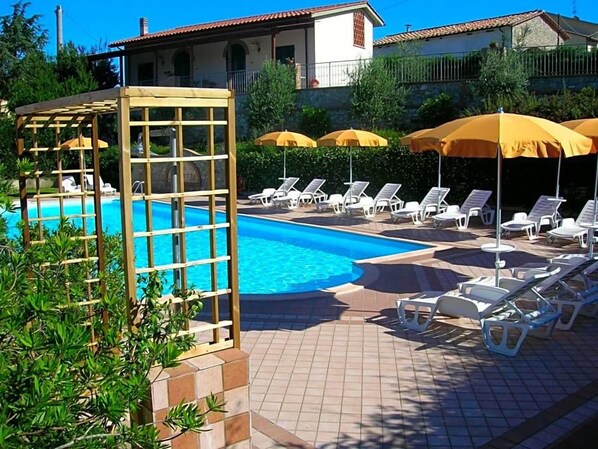 A swimming pool with sun loungers and umbrellas, surrounded by a tiled patio area.