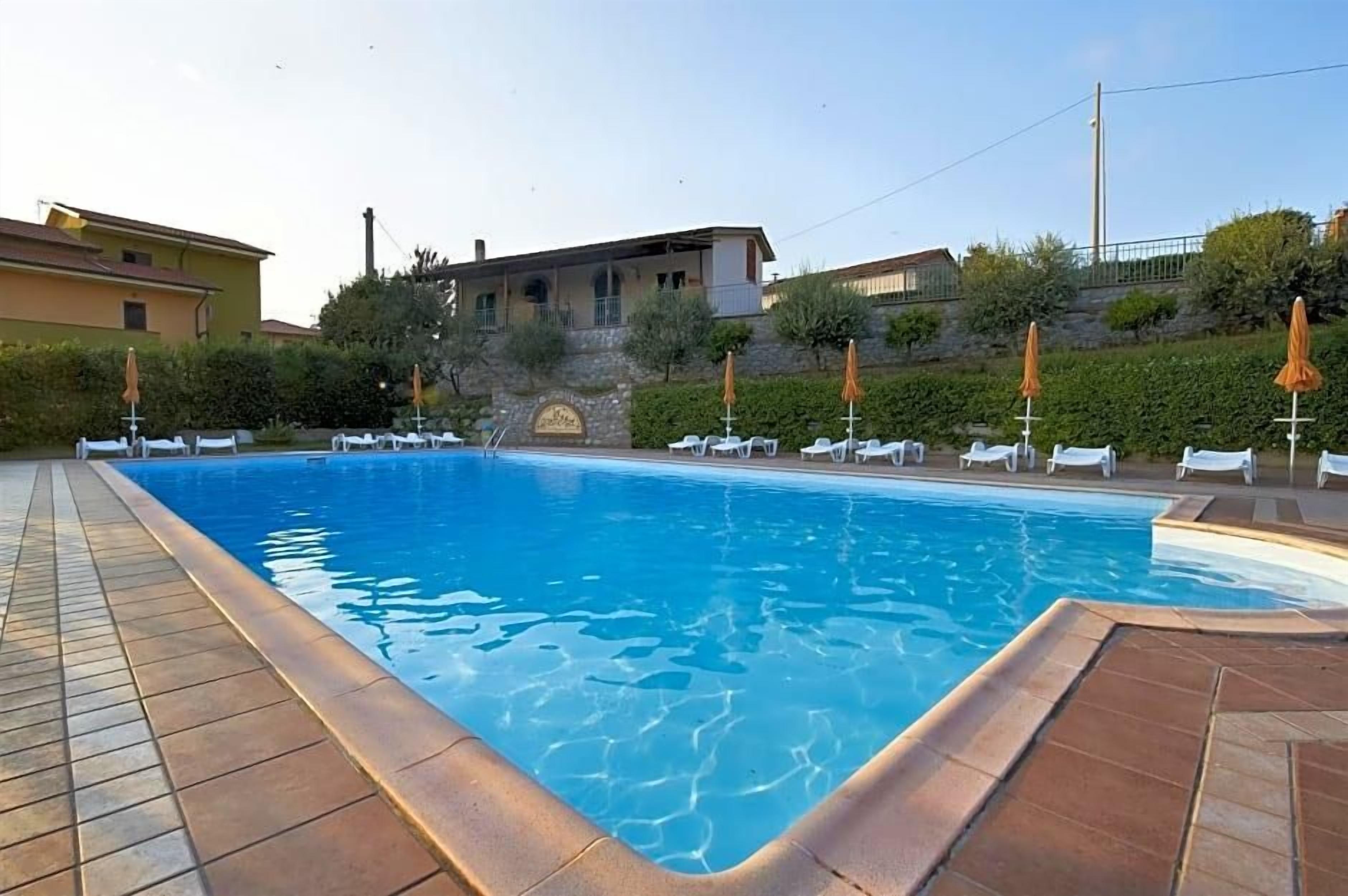 Una piscina con sillones de descanso y sombrillas, rodeada de edificios y follaje.