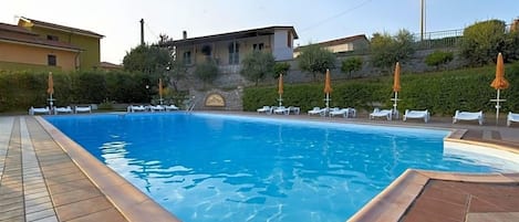 Una piscina con sillones de descanso y sombrillas, rodeada de edificios y follaje.
