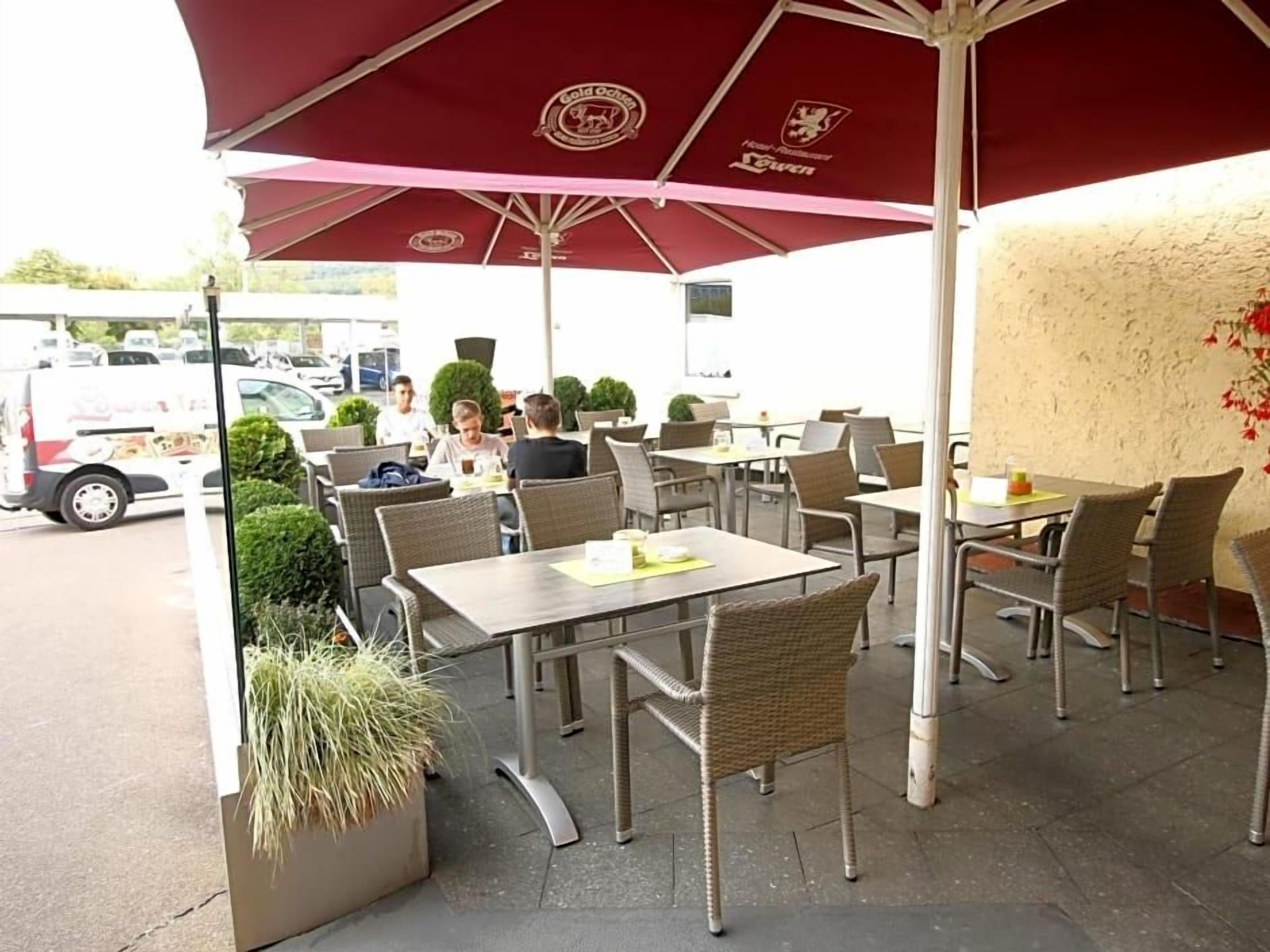An outdoor seating area with tables and chairs, covered by umbrellas, and a white van in the background.