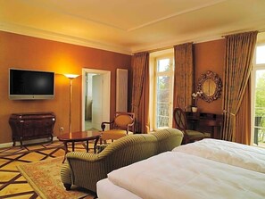 Et hotelværelse med seng, fjernsyn, stol, bord og et vindue med gardiner.