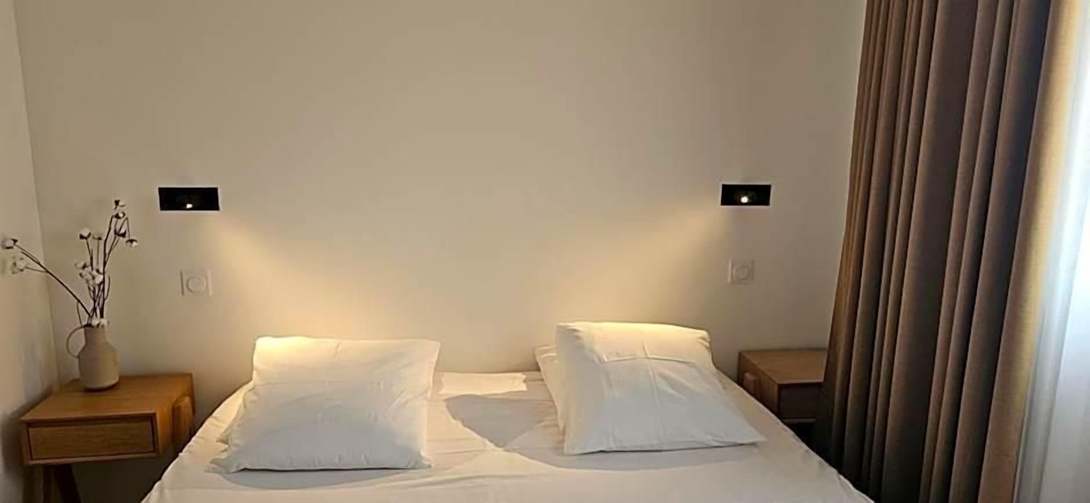 A hotel room with a bed, two bedside tables, a vase with flowers, and a curtain.
