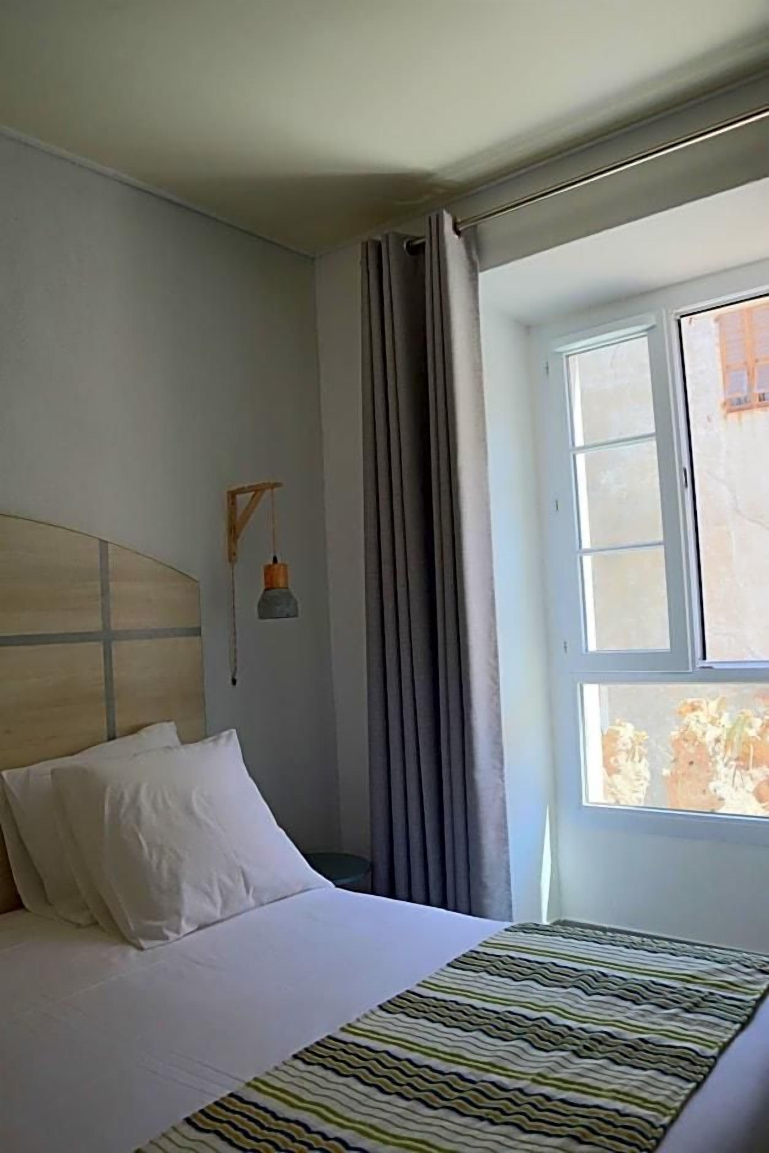 A hotel room with a bed, a window with curtains, a wall-mounted lamp, and a view of the outside.