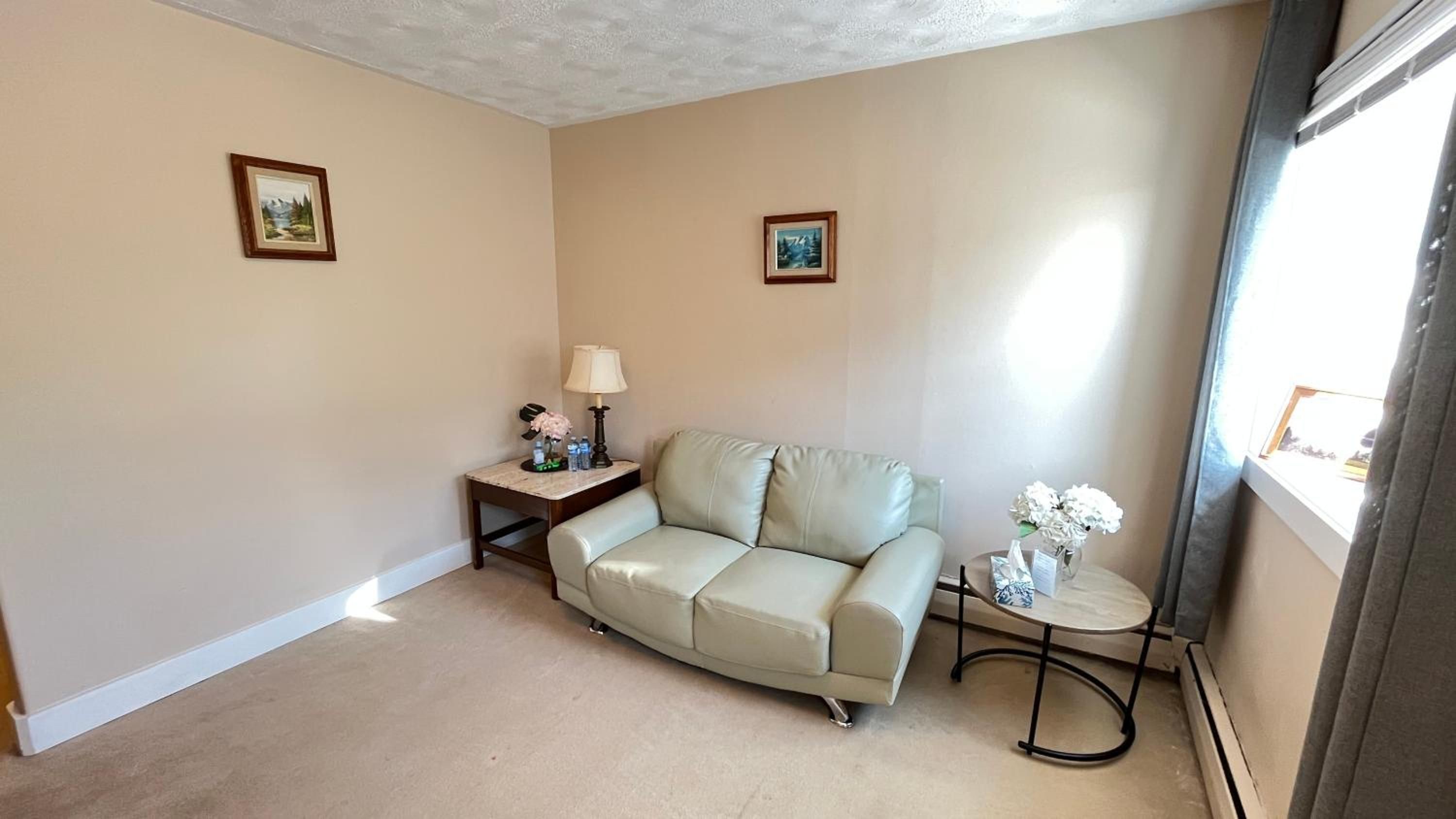 A living room with a beige sofa, a small table with a lamp, and two framed pictures on the wall.