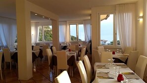 A dining area with tables set for meals, large windows with curtains, and a view of the landscape. - Hotel L'ogliera (Marciana)