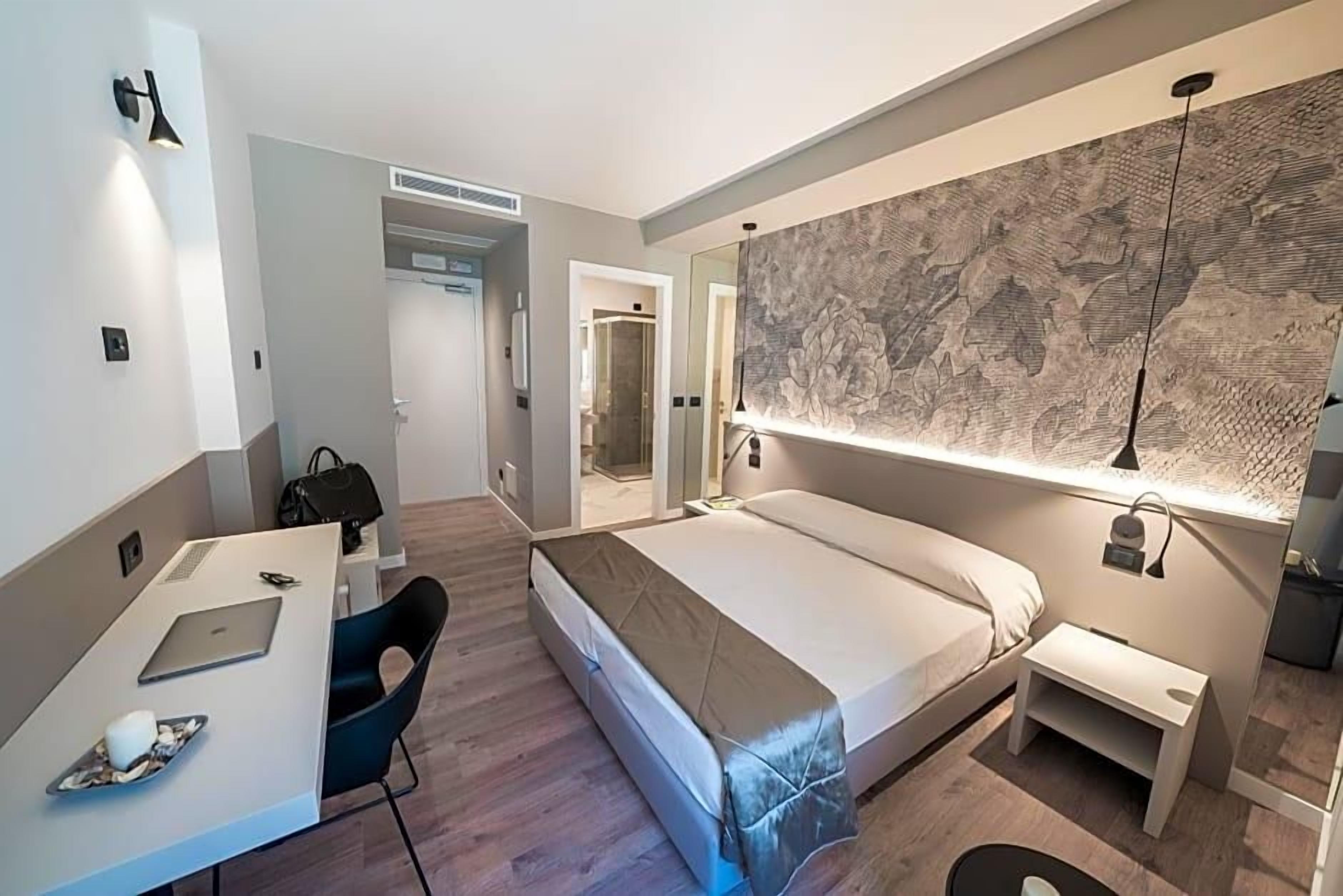 A modern hotel room with a bed, a desk with a laptop, a chair, and a large mirror.