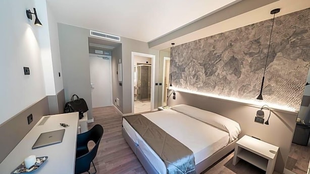 A modern hotel room with a bed, a desk with a laptop, a chair, and a large mirror. - Comfort Zone (Codroipo)