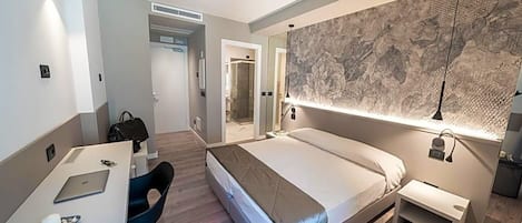 A modern hotel room with a bed, a desk with a laptop, a chair, and a large mirror.