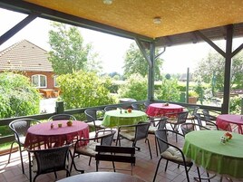 An outdoor patio with round tables and chairs, a covered roof, and a view of a house and greenery.
