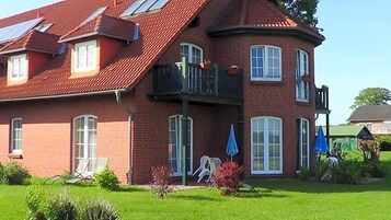 Une maison à deux étages, construite en briques, avec un toit rouge, un balcon et un petit jardin agrémenté d’une chaise longue et d’un parasol bleu.