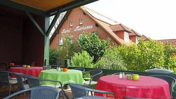 Ein Terrassenbereich im Freien mit Tischen und Stühlen, ein Backsteinhaus mit der Beschilderung 'Hotel Marianne' und Grünflächen im Hintergrund.