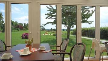 Ein Essbereich mit einem Tisch für zwei Personen, Stühlen und Blick auf einen Garten durch große Fenster.