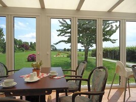 Un coin repas avec une table dressée pour deux personnes, des chaises et une vue sur un jardin grâce à de grandes fenêtres.