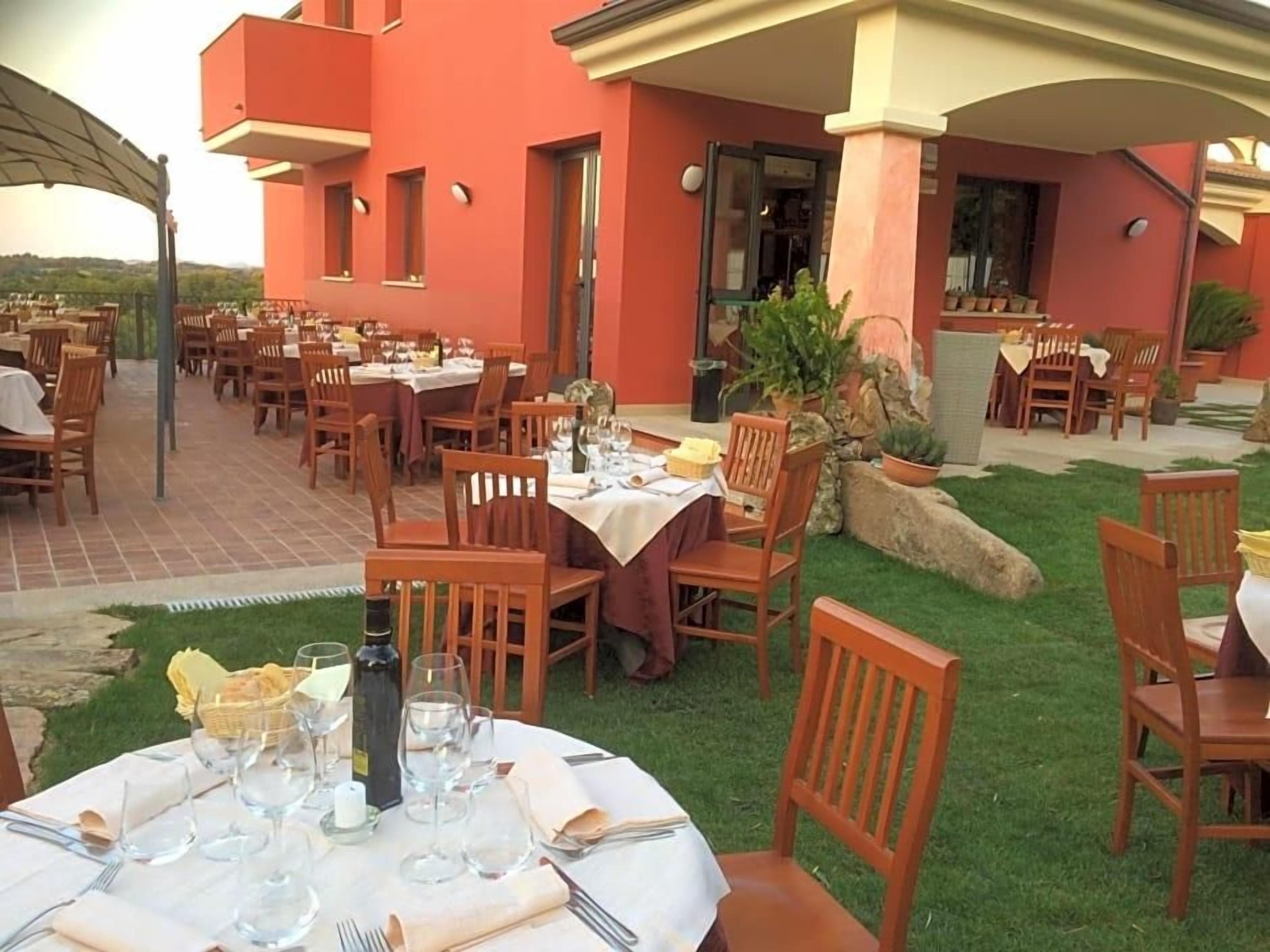 An outdoor dining area with multiple tables and chairs, a building with a red facade, and a grassy lawn.