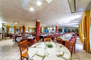 Ampla área de refeições com várias mesas e cadeiras, teto alto com iluminação embutida e um bar ao fundo.