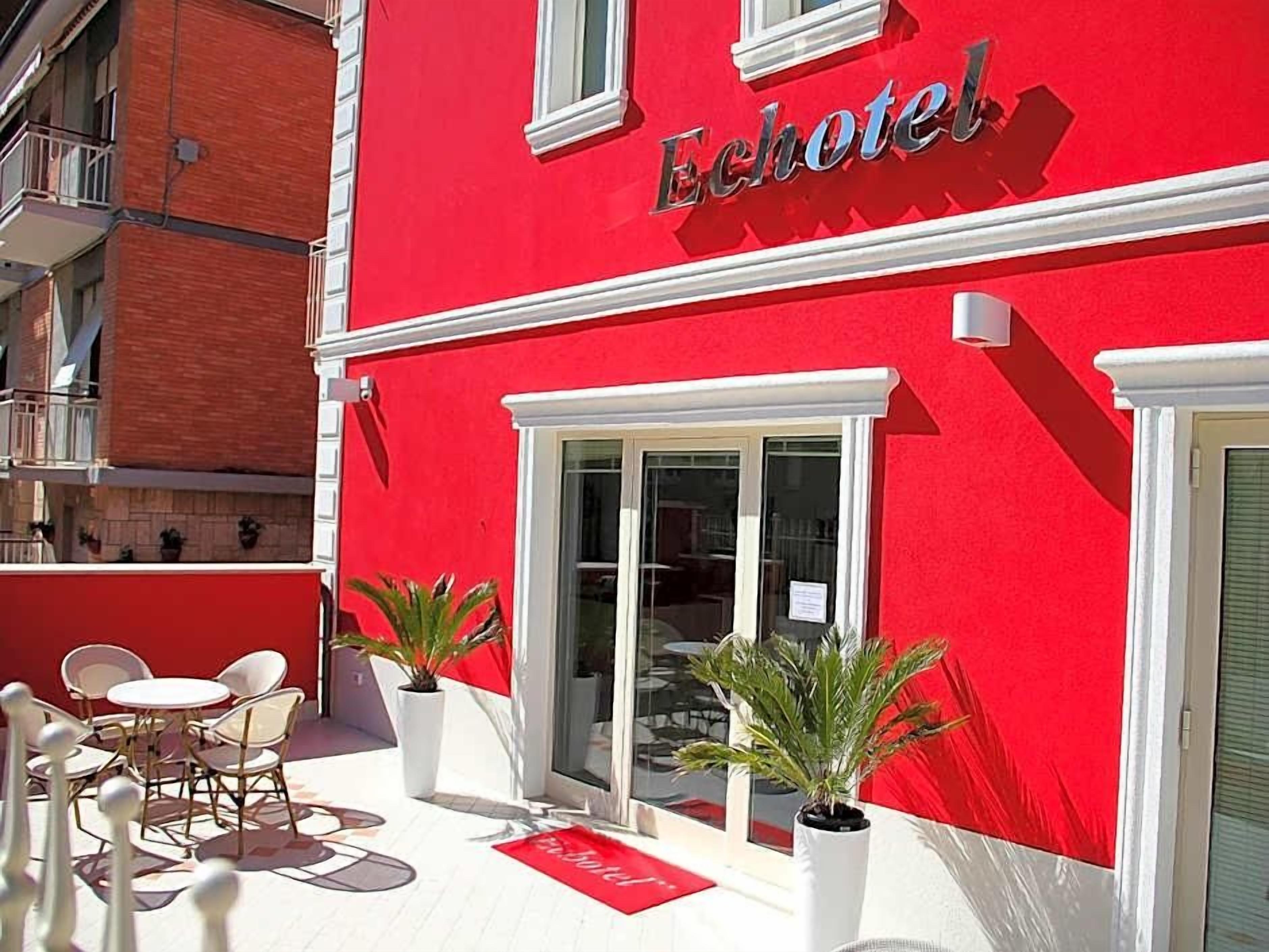 Un edificio rosso con la scritta 'Echo Hotel' sulla facciata. Sono presenti aree salotto all'aperto con sedie e tavoli bianchi.