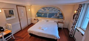 A hotel room with a bed, a desk, a chair, a window with curtains, and a painting on the wall.