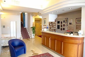 A hotel lobby with a reception desk, a blue chair, a staircase, and a wall with framed pictures. - Hotel Cristallo (Fano)