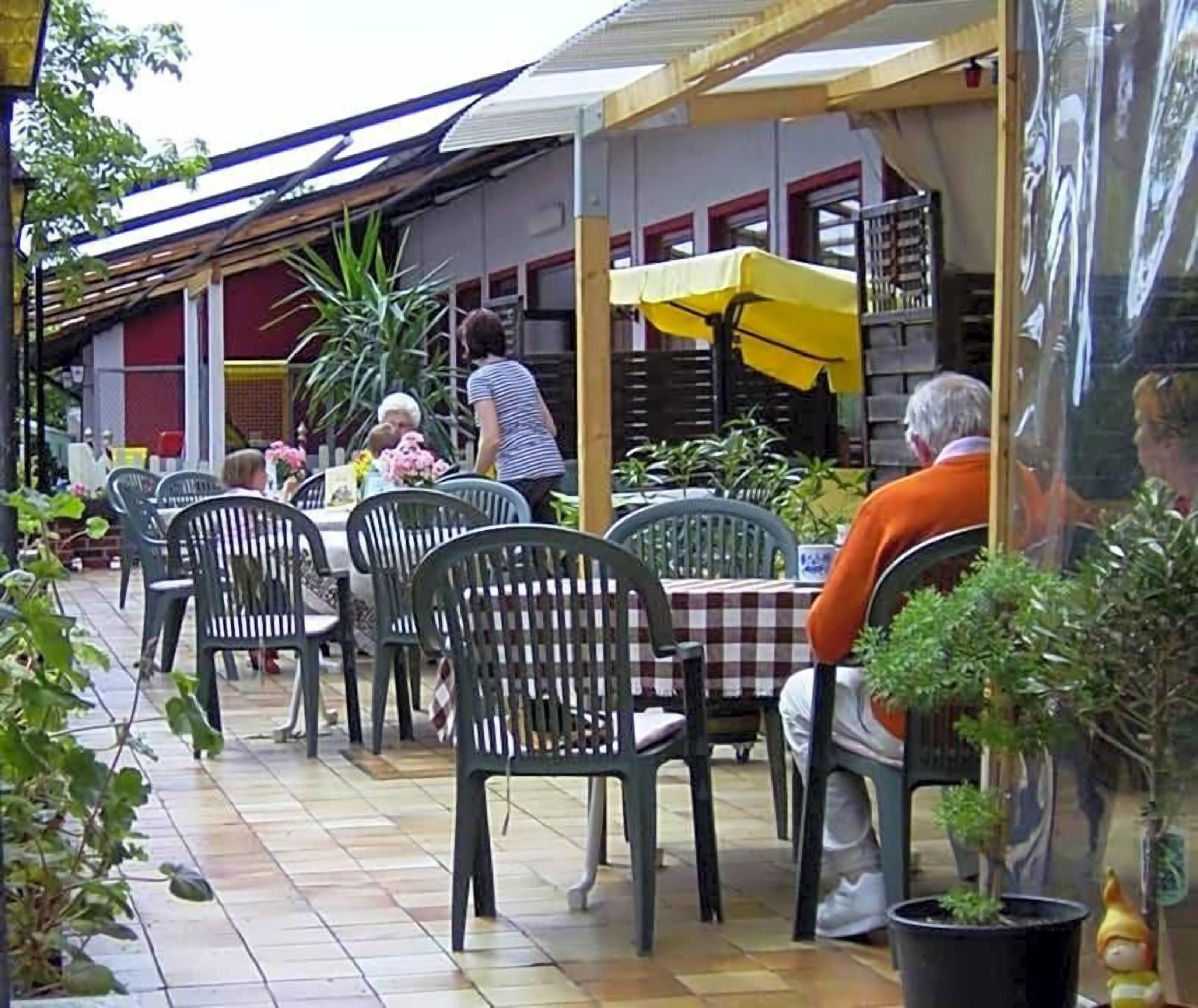 Un cafè a l'aire lliure amb taules i cadires, un para-sol groc i plantes en testos.