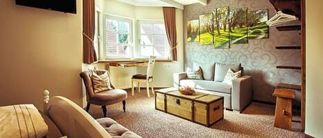 A living room with a sofa, armchair, and a coffee table. There is a window with curtains, a flat-screen TV mounted on the wall, and a painting above the sofa.