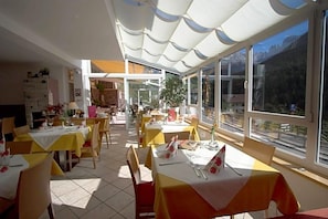 Un restaurante con mesas dispuestas para comer, amplios ventanales que ofrecen vistas al exterior y una zona de estar cubierta al aire libre.