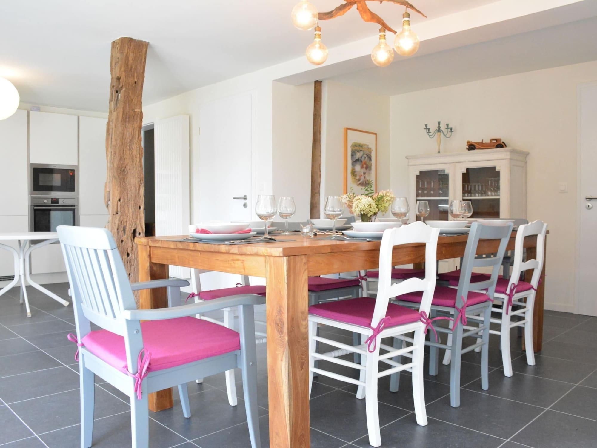 Een eetgedeelte met een houten tafel, witte stoelen met roze kussens en een moderne keuken op de achtergrond.