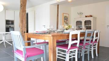 Een eetgedeelte met een houten tafel, witte stoelen met roze kussens en een moderne keuken op de achtergrond.