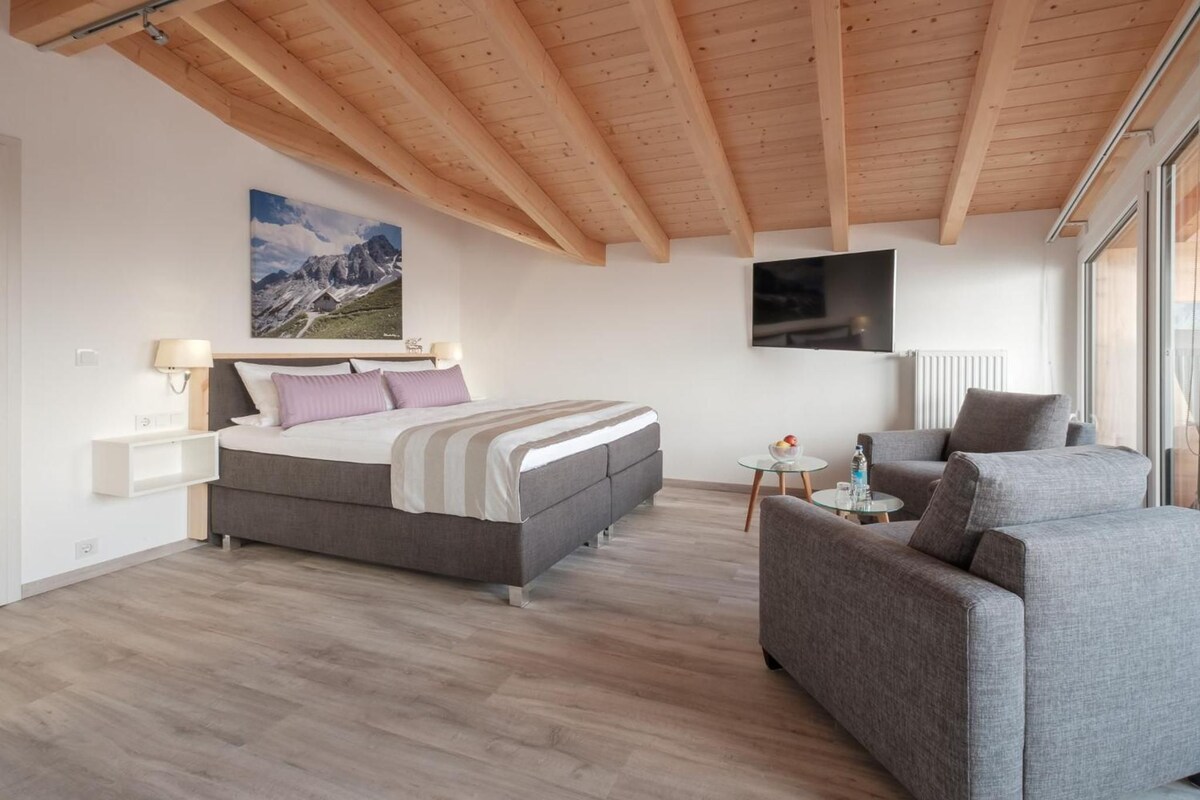 Quarto moderno com cama, sofá, televisão e uma pintura de montanha na parede.