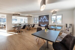 Una habitación de hotel moderna con un ventanal grande, una zona de comedor con mesas y sillas, y un logotipo en la pared.