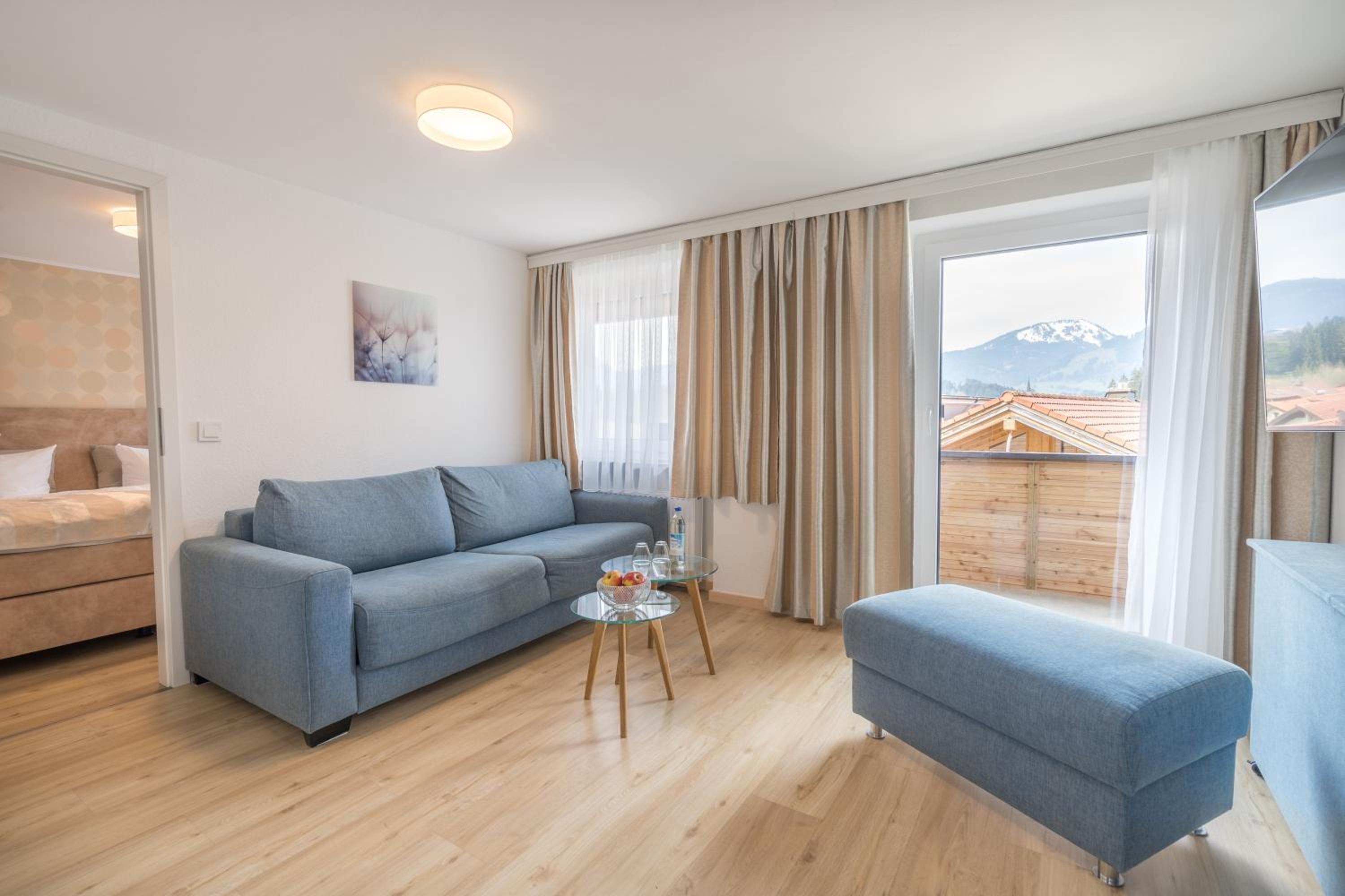 Ein modernes Wohnzimmer mit einer Couch, einem kleinen Tisch mit Gläsern und einer Wasserflasche, und Blick auf ein Gebirge durch große Fenster.