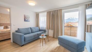Ein modernes Wohnzimmer mit einer Couch, einem kleinen Tisch mit Gläsern und einer Wasserflasche, und Blick auf ein Gebirge durch große Fenster.