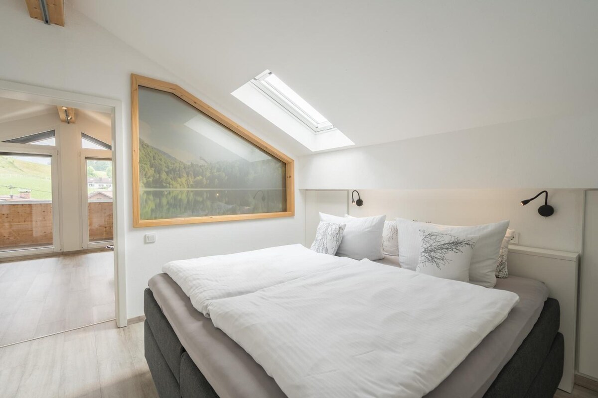 Quarto com claraboia, cama com roupa de cama branca e vista para uma paisagem montanhosa.
