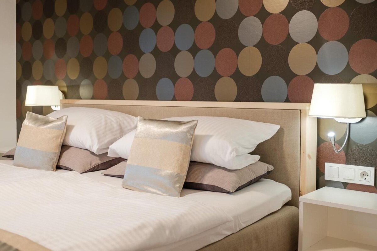 Cama bem arrumada com várias almofadas e duas luminárias de cabeceira.