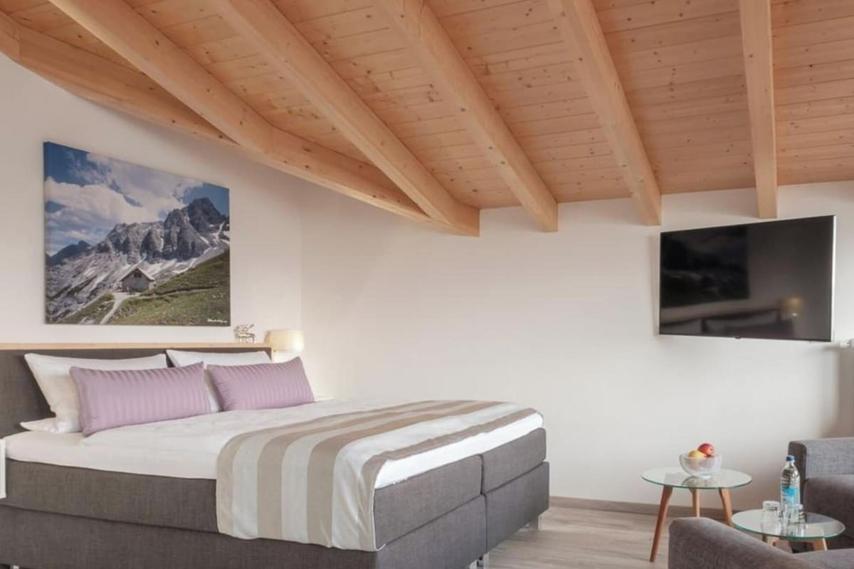 Un dormitorio con cama, una silla, una mesita, un cuadro y un televisor.