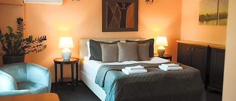 A hotel room with a bed, two bedside lamps, a potted plant, a chair, and a painting on the wall.