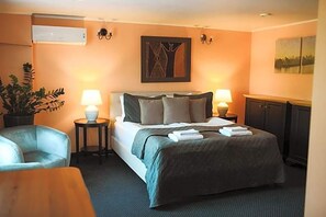 Una camera d'albergo con un letto, due lampade ai comodini, una pianta in vaso, una sedia e un quadro appeso al muro.